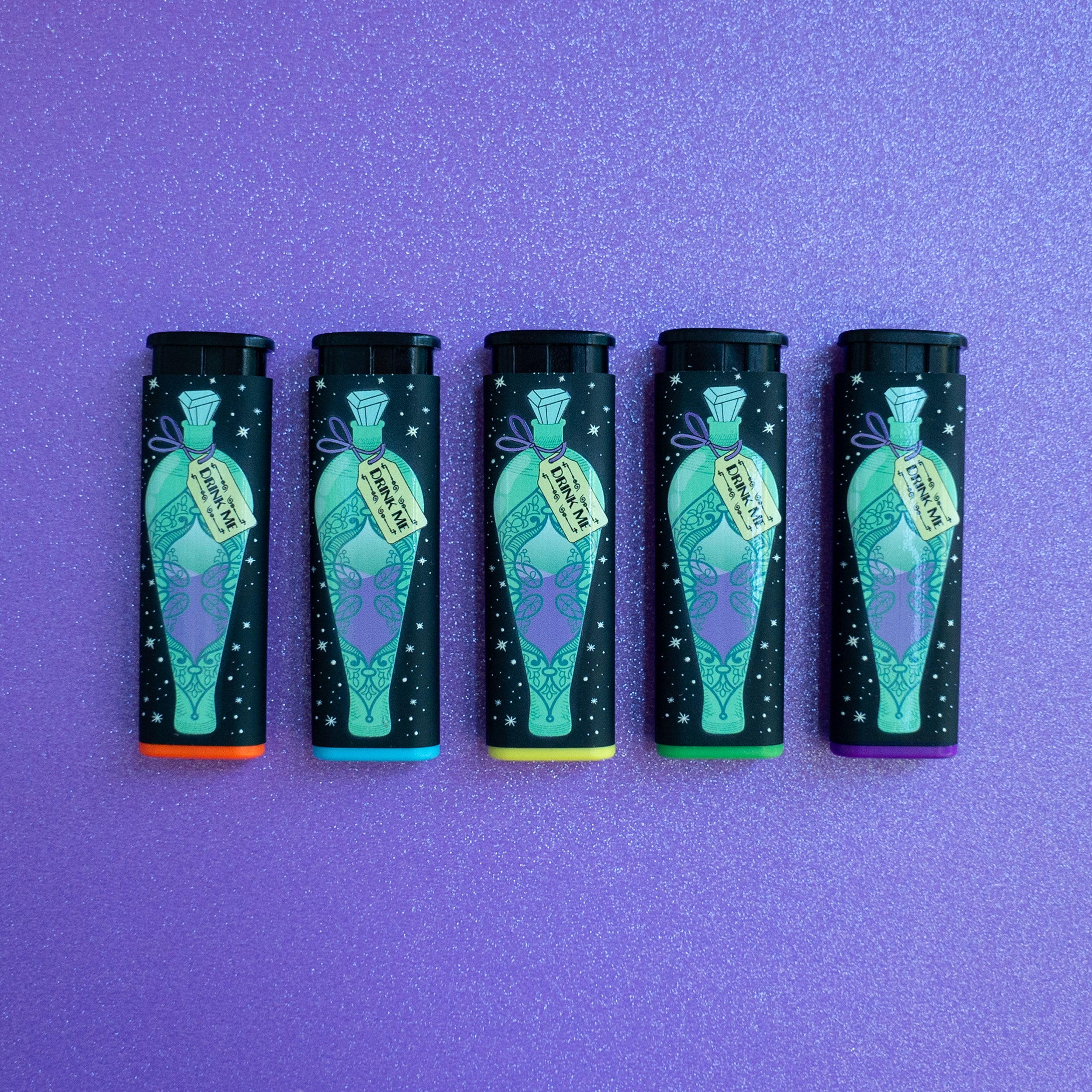 5 Feed Your Head Torch Lighters by Ella Dandelion, showcasing its windproof and refillable design. Featuring the front design, 'Drink Me' green and purple potion. And the back design, green and purple 'eat me' starry illustration. On a light purple glittery background.